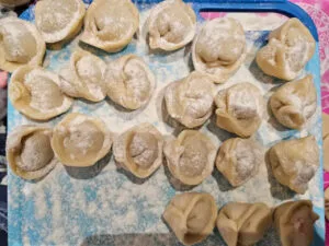 Gotowe, surowe pierogi bezglutenowe oprószone mąką, ułożone na niebieskiej desce. Pierogi z nadzieniem roślinnym