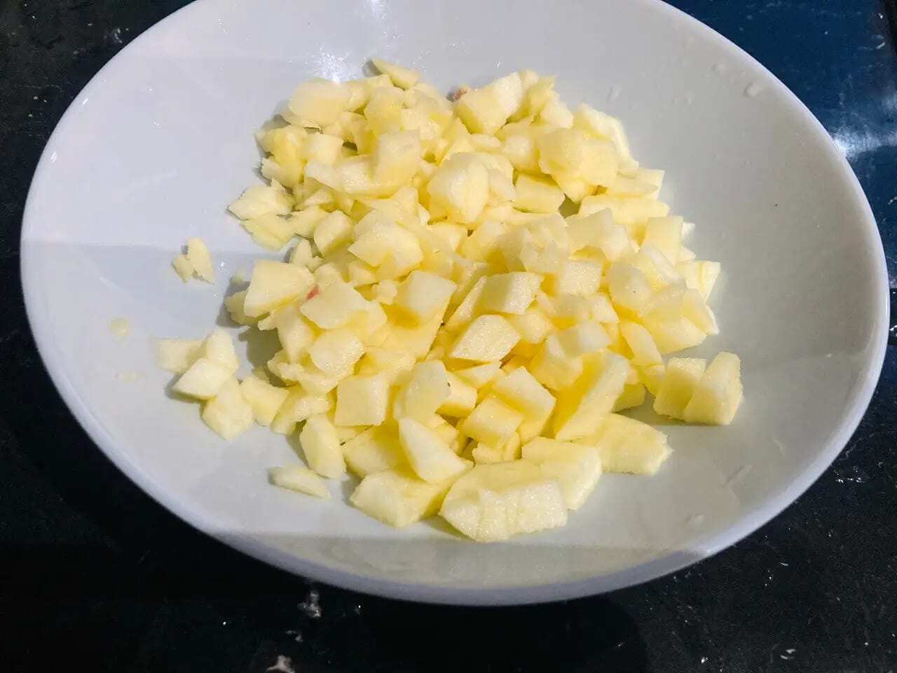 Zbliżenie na miskę pełną świeżo pokrojonych w drobną kostkę jabłek, gotowych do wykorzystania jako nadzienie do domowych pierożków lub szarlotki. Kawałki owoców mają jasnożółty kolor i widoczne są na białym, ceramicznym talerzu, co sugeruje etap przygotowania deseru. Idealne do opisania wizualnej reprezentacji składnika w przepisie kulinarnym na blogu.