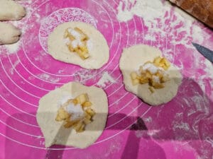 Zbliżenie na trzy surowe pierożki z jabłkami, leżące na jaskraworóżowym, silikonowym blacie kuchennym z nadrukowanymi miarkami. Na otwartym cieście każdego pierożka widoczna jest duża ilość posiekanych jabłek posypanych białym cukrem. Pierogi są gotowe do zamknięcia i dalszej obróbki termicznej. Widoczna mąka rozsypana na blacie