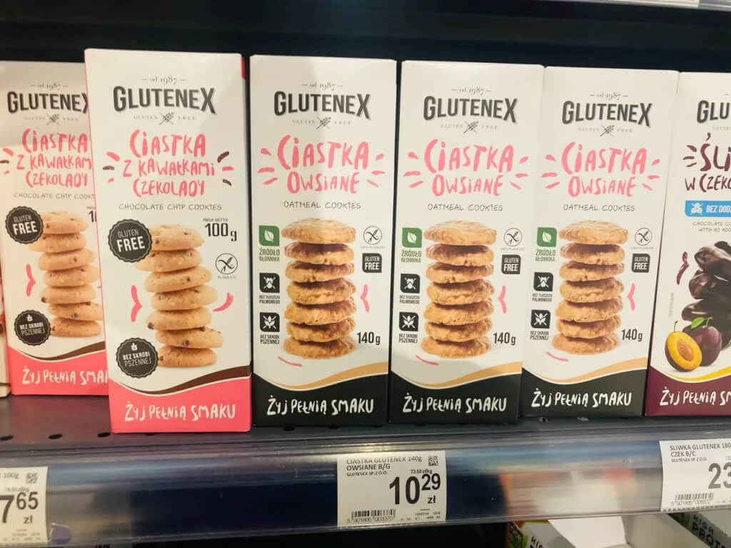 Półka sklepowa z produktami marki Glutenex: bezglutenowe ciasteczka owsiane oraz ciastka z kawałkami czekolady w estetycznych opakowaniach z widocznymi cenami.