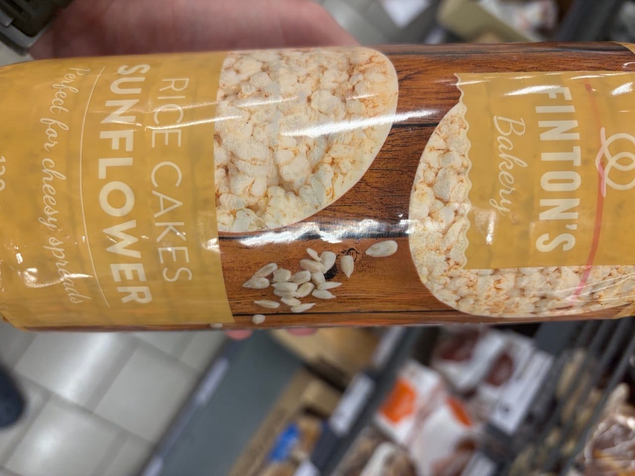 Opakowanie wafli ryżowych ze słonecznikiem marki Finton's Bakery z napisami Rice Cakes Sunflower oraz Perfect for cheesy spreads na tle imitującym drewno, z widocznym okrągłym waflem i ziarnami słonecznika.