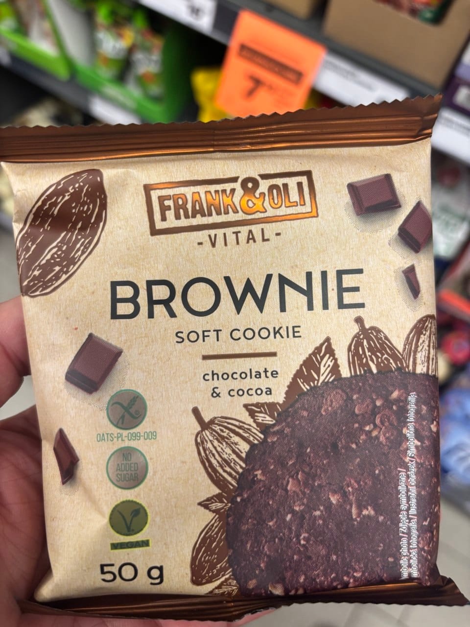 Apetyczne, utrzymane w czekoladowo-kremowej kolorystyce opakowanie miękkiego ciastka Frank & Oli Vital Brownie o wadze 50 g. Na folii, imitującej naturalny papier, rozsypano kostki ciemnej czekolady та ilustracje ziaren kakaowca, które otaczają grafikę mocno czekoladowego, wilgotnego wypieku. W lewym rogu wyraźnie odznaczają się zielone ikony certyfikatu wegańskiego oraz Przekreślonego Kłosa.