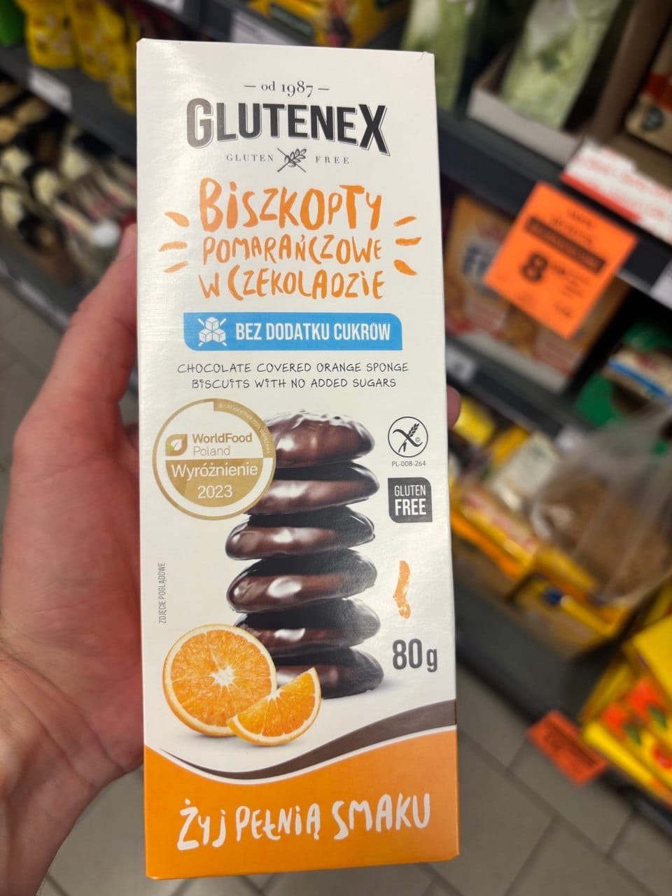 Pionowe opakowanie biszkoptów pomarańczowych w czekoladzie Glutenex Bez dodatku cukrów, z grafiką stosu ciastek, plastrów pomarańczy oraz znakiem Przekreślonego Kłosa.
