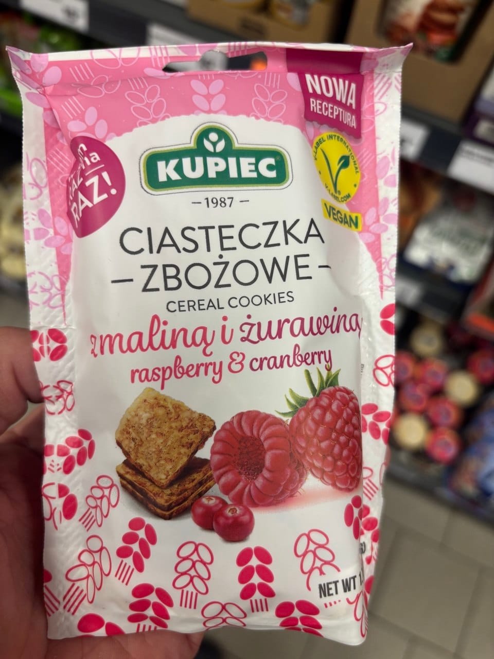 Biało-różowe opakowanie ciasteczek zbożowych Kupiec z maliną i żurawiną, z grafiką owoców, ciastek oraz żółtym certyfikatem Vegan.