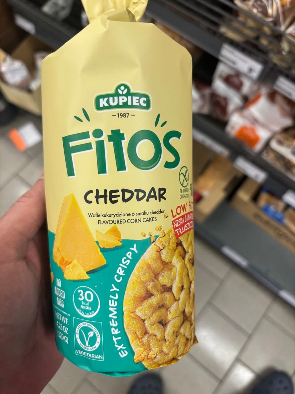 Opakowanie wafli kukurydzianych Kupiec Fitos o smaku Cheddar, na którym widnieje duże zielone logo marki, kawałki żółtego sera oraz chrupiące krążki kukurydziane z napisem Extremely Crispy
