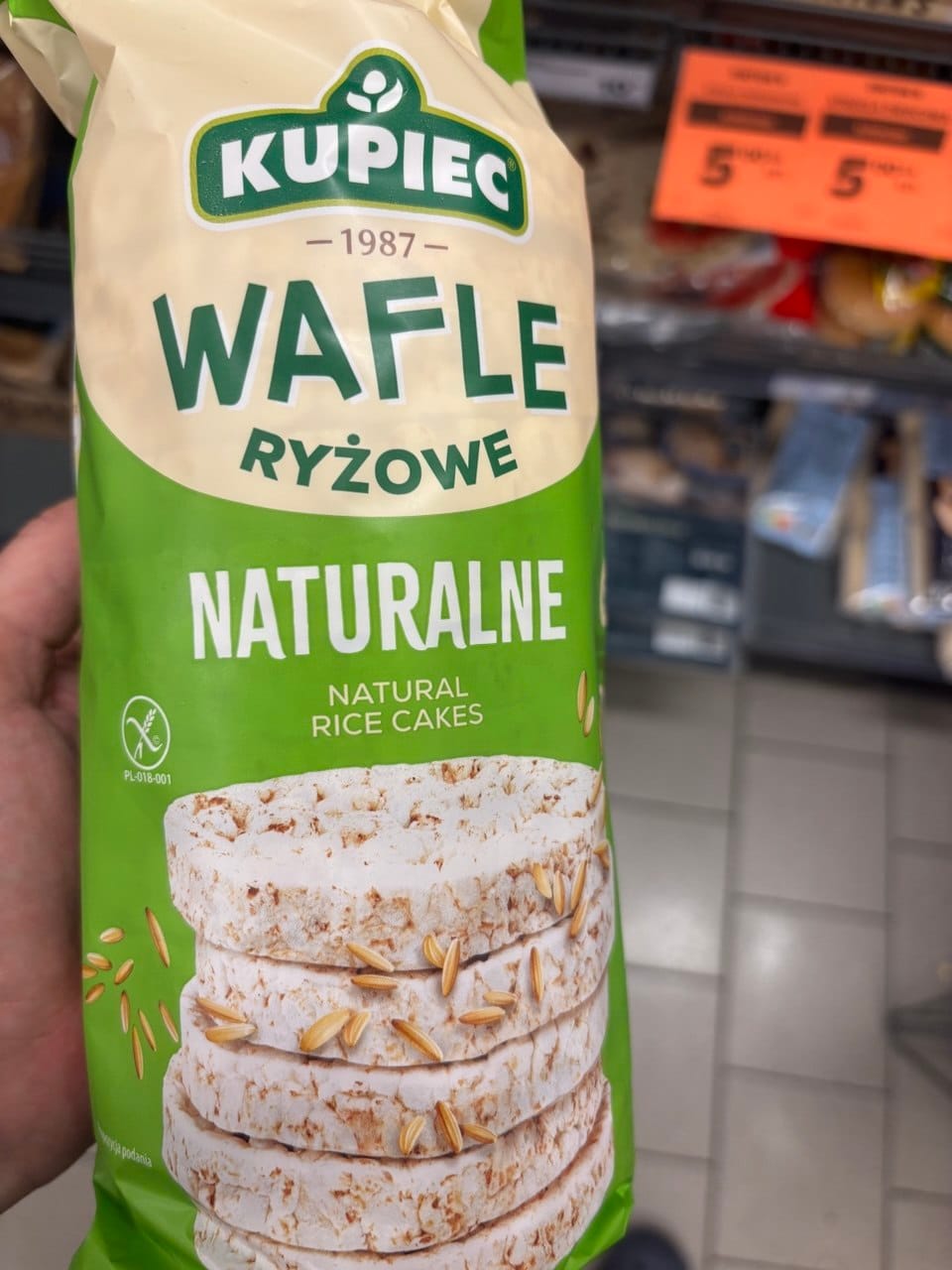 Zielono-białe opakowanie wafli ryżowych Kupiec Wafle Ryżowe Naturalne z widocznym symbolem Przekreślonego Kłosa, logotypem marki oraz grafiką przedstawiającą stos chrupiących wafli z ziarnami ryżu.
