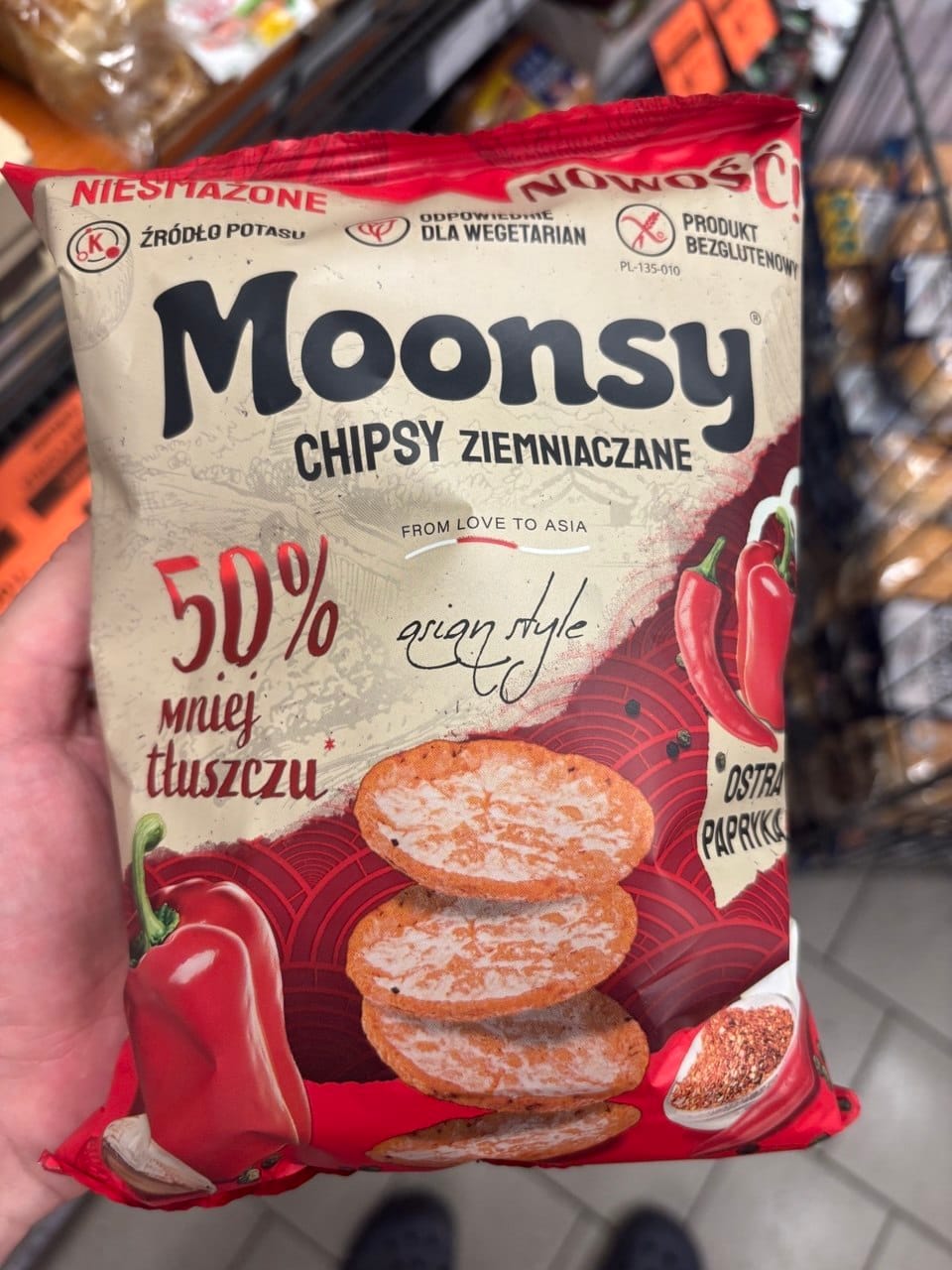 Opakowanie chipsów ziemniaczanych Moonsy o smaku Ostra Papryka z napisem 50% mniej tłuszczu, Niesmażone oraz From Love to Asia, przedstawiające okrągłe chrupki, papryczki chili i symbol Przekreślonego Kłosa oznaczający produkt bezglutenowy.