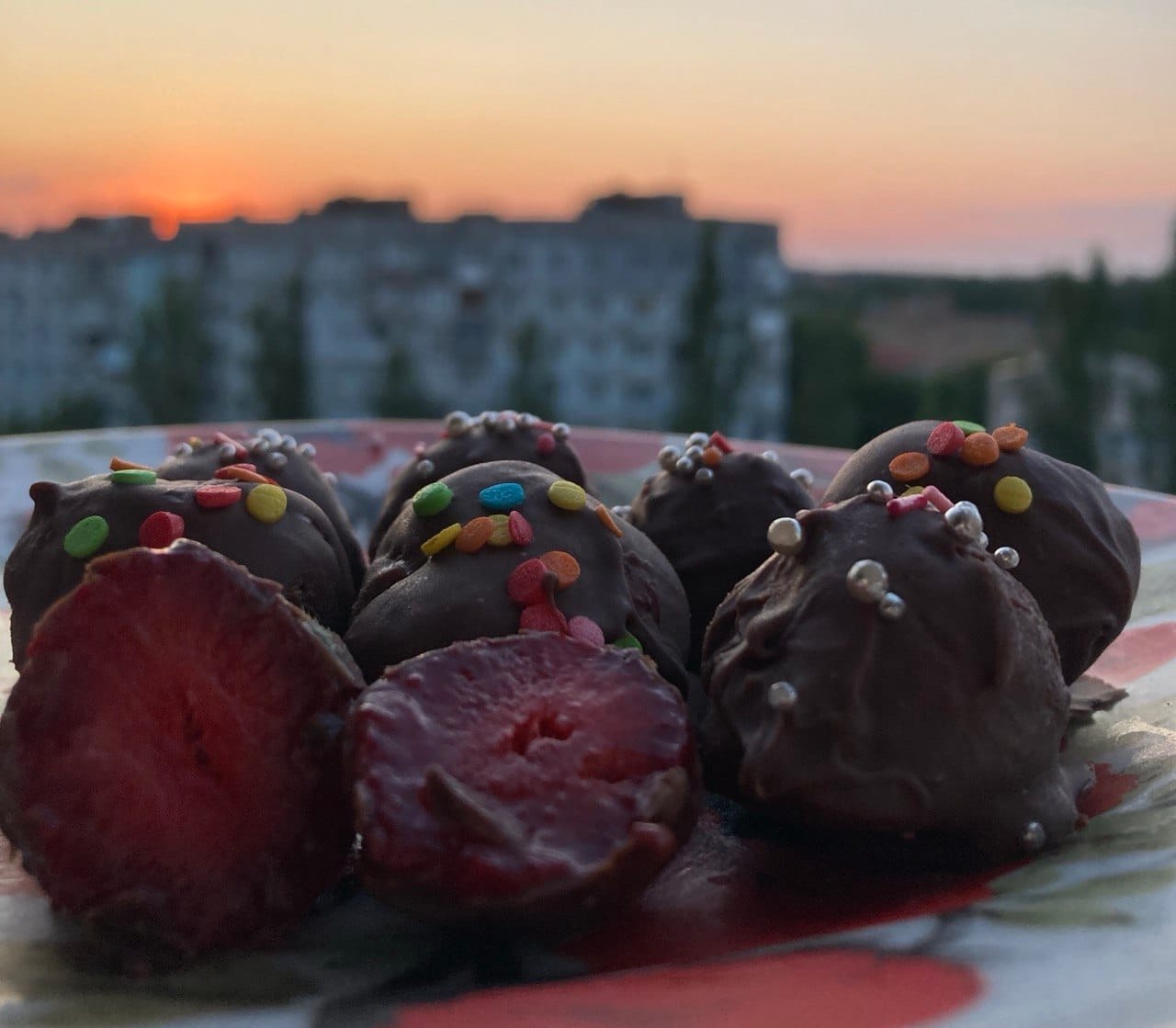 Fresh strawberries covered in dairy-free chocolate with colorful sprinkles, served on a plate against a city sunset skyline in Armenia.