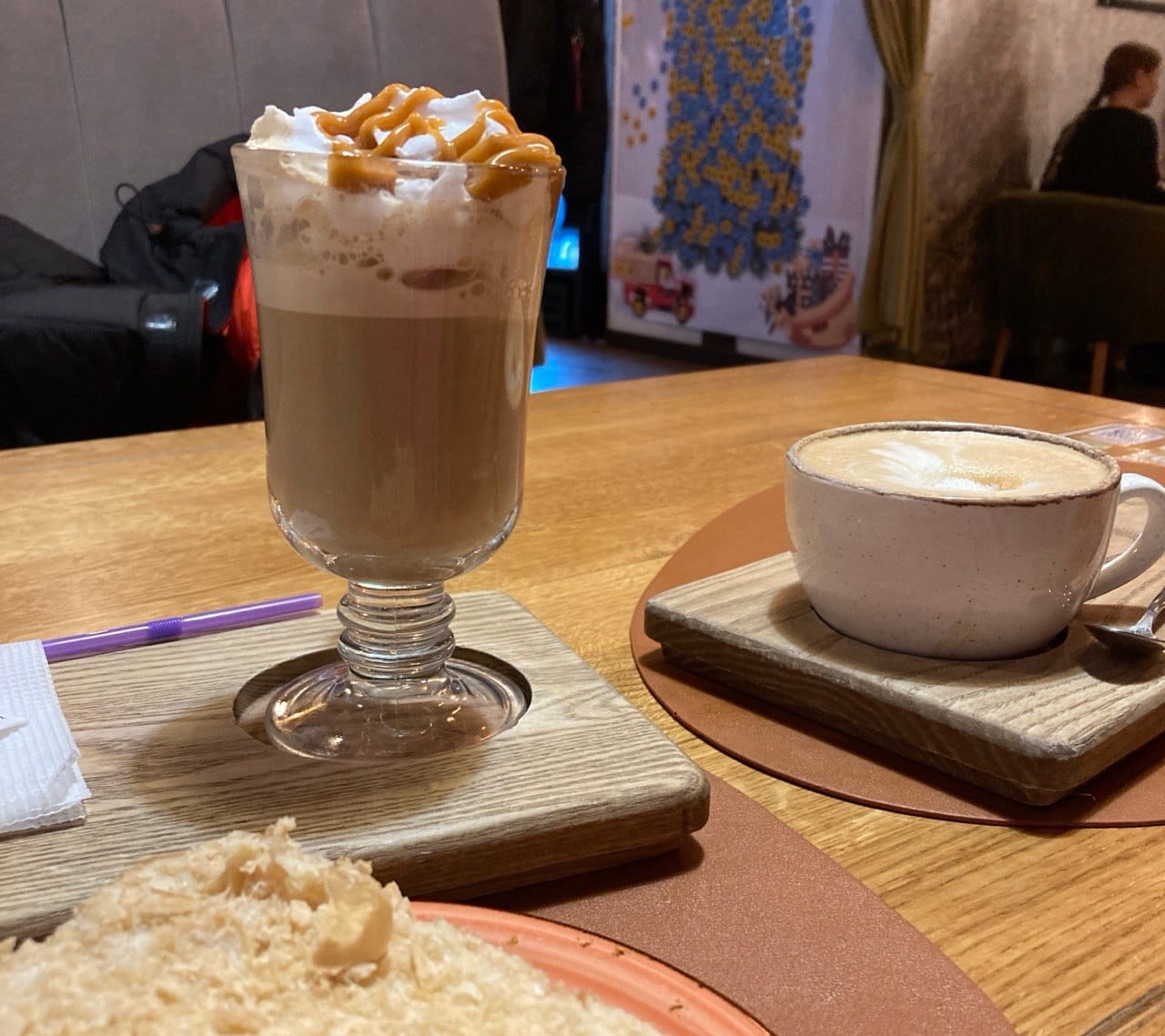 Two coffee drinks on a wooden table in a cozy Baltic café: a tall glass with a creamy caramel-topped beverage and a classic cappuccino in a white ceramic cup, next to a fluffy sponge dessert. This image captures the cafe atmosphere explored in the article comparing gluten-free travel and celiac-friendly dining options in Vilnius and Riga.
