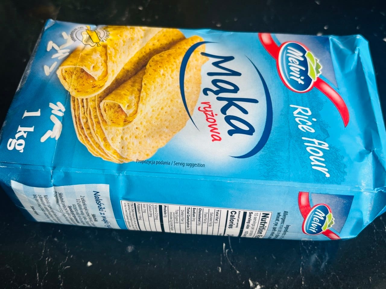 A blue 1 kg package of Melvit white rice flour resting on a dark surface